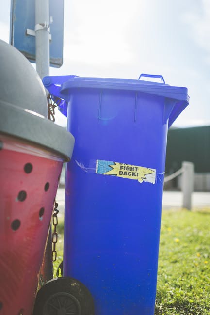 A large green waste disposal bin filled with a mixture of cardboard boxes, some flattened and others in a pile, alongside white packaging cartons and light wooden pallets leaning against the side. The