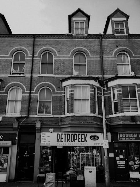 A black-and-white photograph of a multi-storey brick building with large, arched windows on the first and second floors, and smaller dormer windows on the top floor. The building features a protruding